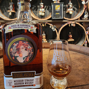 Missouri Straight Bourbon Whiskey bottle with a glass on a wooden surface, with decorative barrels in the background.