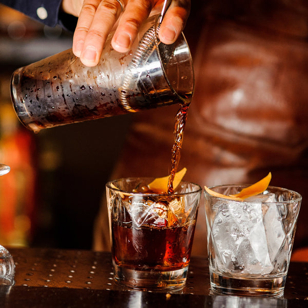  person pouring a cocktail from a shaker into a glass filled with ice cubes on a bar counter, with another glass partially visible in the foreground.